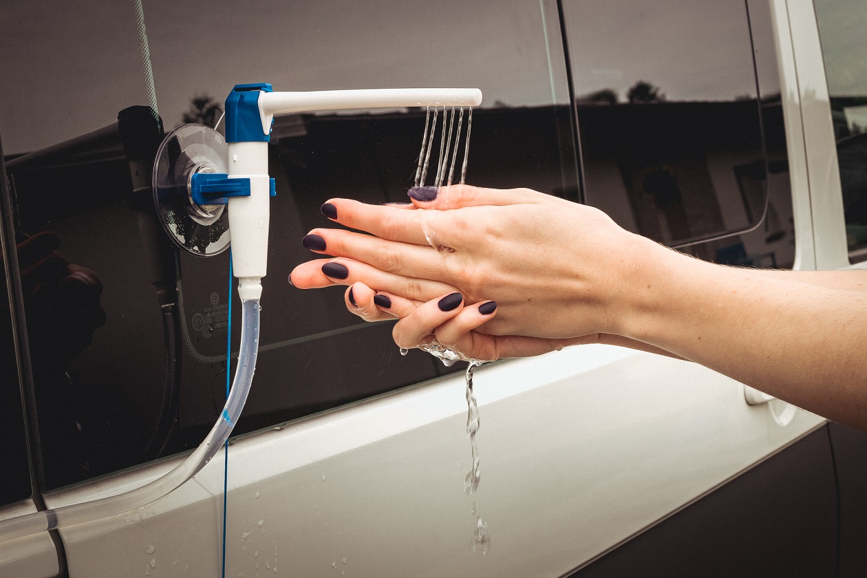 Zbliżenie na kobiece dłonie oraz przyczepiony do białej karoserii za pomocą przyssawek kran, z którego leci woda na kobiece dłonie